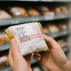 コンビニの陳列棚前で栄養成分表示を確認する手元、糖類・飽和脂肪酸・食塩相当量に赤線で目線が向くイメージ。