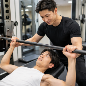 パーソナルジムでトレーナーとマンツーマン指導を受けながら筋トレに取り組む痩せ型の日本人男性